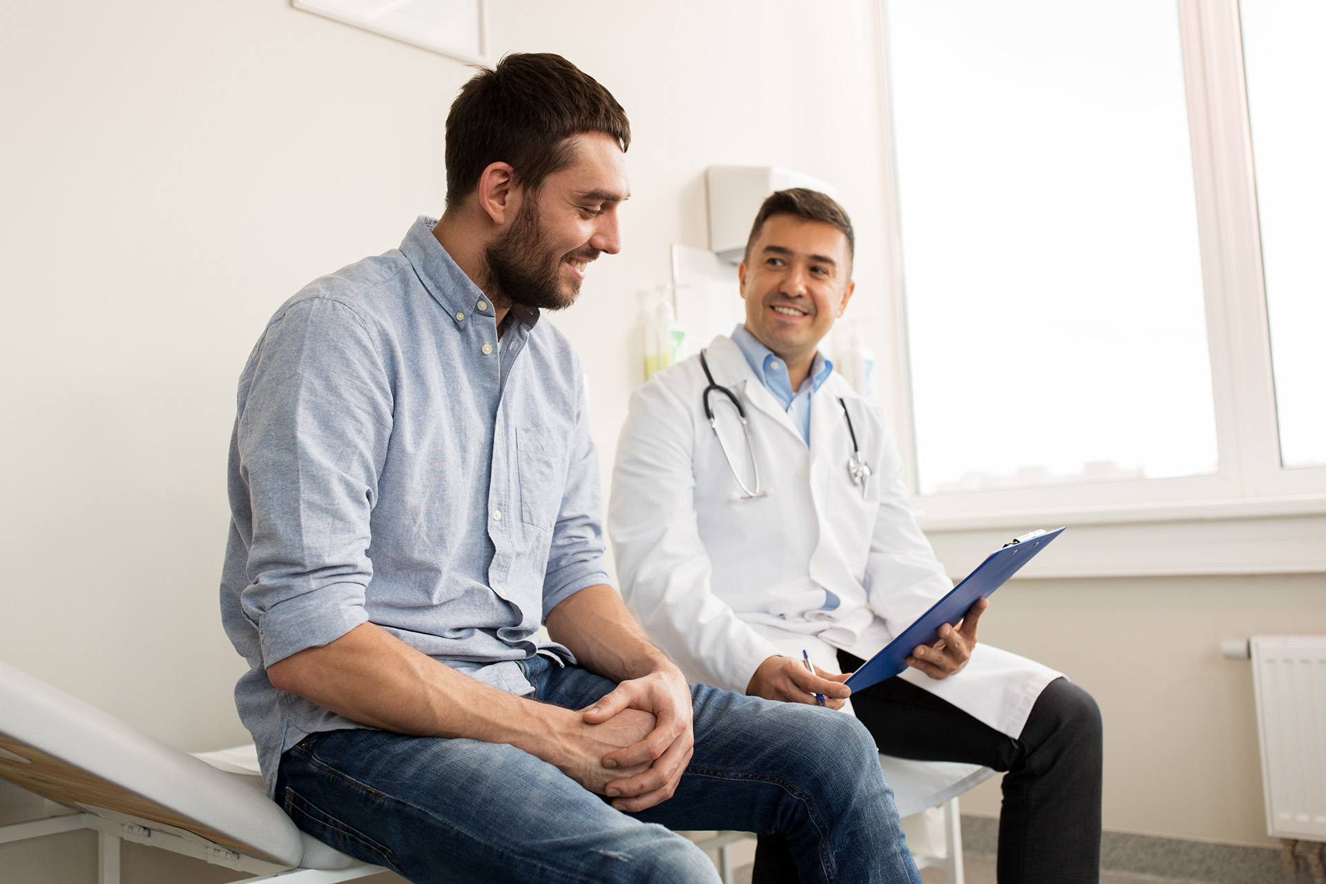 medicine, healthcare and people concept - smiling doctor with clipboard and young man patient meeting at hospital